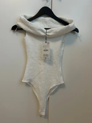 Vit body med krage från ZARA ny med prislapp - Säljer en vit body från ZARA i storlek S. Bodyn har en bred, uppvikt krage och är tillverkad i ett mjukt, frottéliknande material. Perfekt för dig som vill ha en stilren och bekväm look. Snygg passform och enkel att matcha med andra plagg.
