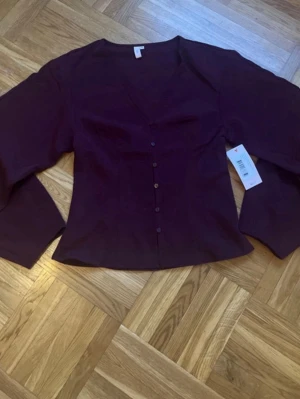 Vinröd blus med knappar - Stilren vinröd blus med v-ringning och knappar framtill. Blusen har långa ärmar och lätt figursydd passform. Perfekt för dig som gillar en enkel men elegant look. Materialet är mjukt och följsamt.