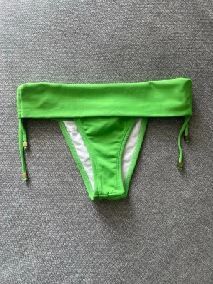 Gröna bikiniunderdelar med knyt från Brasilien - Snygga gröna bikiniunderdelar från Brasilien  med bred linning och knyt i sidorna med gulddetaljer. Modellen har en enkel och stilren design, perfekt för strand eller pool. Insidan är vitfodrad för extra komfort. #bikini #brasilien 