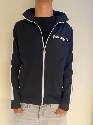 Palm Angels svart ziptröja S - Svart ziptröja från Palm Angels med vit dragkedja och vita ränder längs ärmarna. Hög krage och logga på bröstet. Perfekt för en streetwear-look och enkel att matcha med jeans eller joggers.