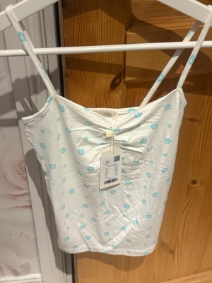 Vit pyjamas med blå blommor - Söt pyjamas med vitt linne och matchande byxor, båda med små blå blommor. Linnet har tunna axelband och en liten rynkning framtill. Byxorna har resår i midjan och avslappnad passform. Perfekt för mysiga kvällar hemma.