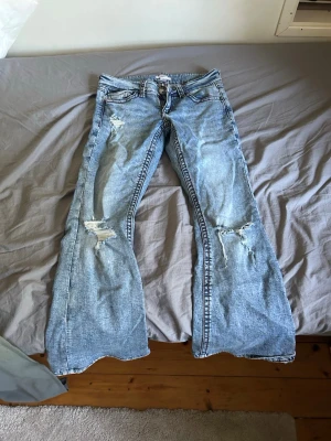 Ljusblå bootcut jeans med slitningar - Säljer ett par ljusblå bootcut jeans med slitna detaljer på både fram- och baksida. Jeansen har klassiska fem fickor och markerade sömmar. Perfekta för en avslappnad och trendig look. Materialet är denim i bomull.