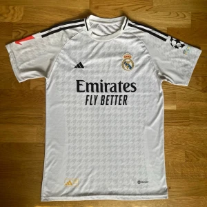 Real Madrid matchtröja Bellingham - Säljer en vit Real Madrid matchtröja från Adidas med Bellingham och nummer 5 på ryggen. Tröjan har svarta detaljer, klubbmärke på bröstet, Champions League-märke på ärmen och sponsortryck framtill. Tillverkad i lätt och ventilerande material.