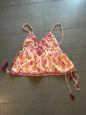 Mönstrad linnetopp från Design by Amor - Säljer en färgglad linnetopp från Design by Amor i storlek XS. Toppen har tunna axelband, broderade detaljer, snörning framtill och ett orange och rosa mönster på vit botten. Perfekt för varma dagar och har söta tofsar i sidorna.