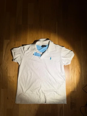 Vit pikétröja från Ralph Lauren - Vit pikétröja från Ralph Lauren med klassisk krage och korta ärmar. Tröjan har en ljusblå logga broderad på bröstet och ljusblå detaljer på insidan av kragen. Perfekt för en stilren och sportig look.