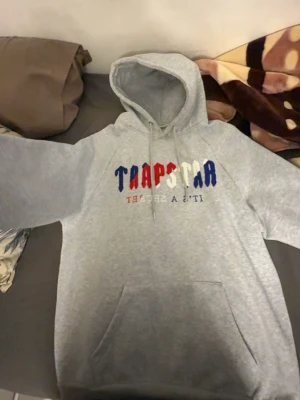 Grå Trapstar hoodie med tryck - Säljer en grå hoodie från Trapstar med färgglatt tryck i blått, rött och vitt på bröstet. Hoodien har snörning i huvan och en stor magficka. Perfekt för dig som vill ha en chill och stilren look.