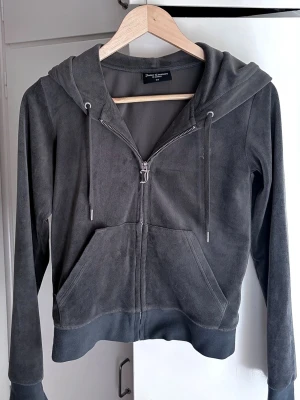 Juicy Couture Hoodie - Mörkgrå hoodie i mjukt velourmaterial med dragkedja framtill och två stora fickor. Modellen har huva med dragsko och ribbade muddar vid ärmslut och nederkant. Perfekt för en avslappnad och trendig look.