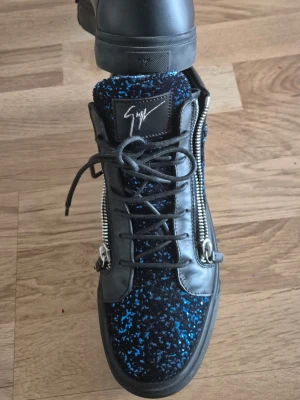 Svarta sneakers med blått mönster - Sälsynta svarta sneakers från Giuseppe Zanotti med blått spräckligt mönster på ovandelen, dubbla silverdragkedjor på sidorna och svart läderdetaljer. Skorna har en hög plös med logga och svarta skosnören. Perfekt för dig som vill sticka ut. Storlek 44 passar även 45.