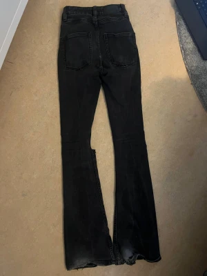 Svarta utsvängda jeans med hål - Säljer ett par svarta jeans med utsvängda ben och råa kanter nertill. Jeansen har stora hål vid knäna och klassiska bakfickor. Perfekta för dig som gillar en edgy och avslappnad stil.