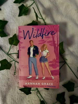 Wildfire av Hannah Grace - Wildfire är en ungdomsroman av Hannah Grace med ett färgstarkt rosa omslag. Boken har en tecknad illustration av två unga vuxna framför ett rosa landskap. Perfekt för dig som gillar romantik och sommarvibbar.