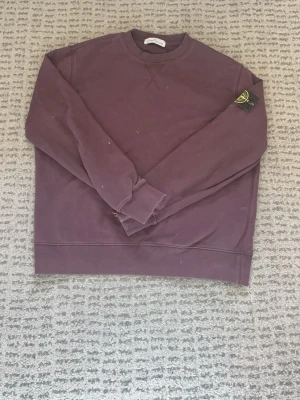 Lila sweatshirt från Stone Island - En stilren lila sweatshirt från Stone Island med klassisk rund hals och ribbade muddar. Tröjan har den ikoniska Stone Island-loggan på vänster ärm och är tillverkad i mjukt bomullsmaterial. Perfekt för en avslappnad och trendig look.