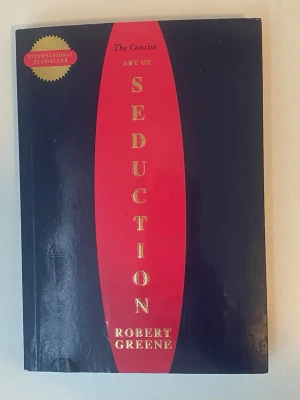 The Concise Art of Seduction - En internationell bästsäljare av Robert Greene. Omslaget är svart med en röd vertikal mitt och guldfärgad text. Boken är i pocketformat och har en modern, stilren design. Perfekt för dig som gillar psykologiska och strategiska teman inom skönlitteratur.