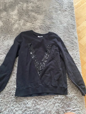 Svart sweatshirt med paljett-V - Säljer en svart sweatshirt med ett stort V i svarta paljetter på bröstet. Tröjan har rund halsringning, långa ärmar och ribbade muddar vid ärmslut och nederkant. Perfekt för dig som gillar en enkel men ändå unik stil.