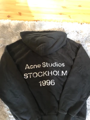 Svart hoodie Acne Studios Stockholm - Svart hoodie från Acne Studios med stor huva och tryck på ryggen där det står 'Acne Studios STOCKHOLM 1996' i vitt. Hoodien har en avslappnad passform och är tillverkad i mjukt material, perfekt för en cool och enkel streetwear-look.