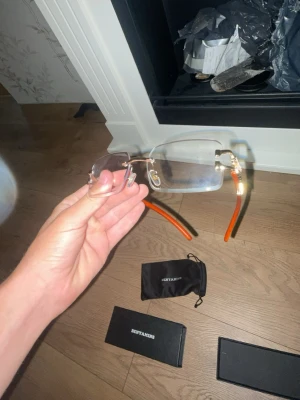 Transparenta solglasögon från Benyamins - Säljer ett par stilrena solglasögon från Benyamins med transparenta glas och guldfärgade detaljer. Skalmarna är orangea och ger en unik touch till looken. Kommer med originalfodral och förvaringspåse.