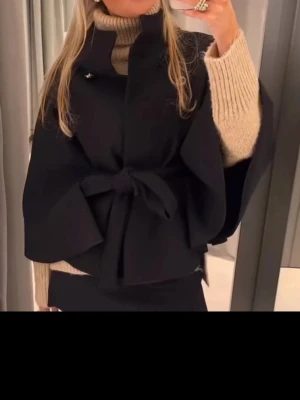 Svart missay kappa - Stilren svart cape-kappa med knytband i midjan och hög krage. Kappan har vida ärmar och en elegant, lös passform som ger en trendig siluett. Perfekt att bära över en stickad tröja för en chic look.