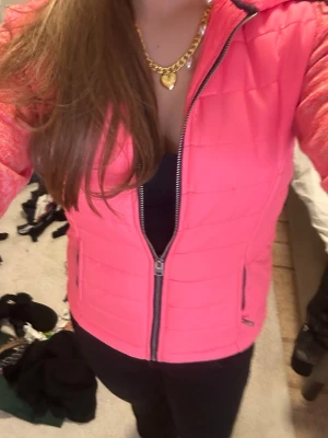 Rosa vindjacka från Superdry - Snygg rosa vindjacka från Superdry med huva och dragkedja framtill. Jackan har quiltad framsida, stickade ärmar i melerad rosa ton och detaljerade logotyper på ärmen. Perfekt för blåsiga dagar och ger en sportig look.