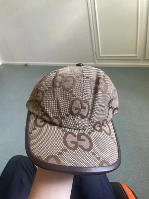Brun keps med GG-mönster - Säljer en brun keps med klassiskt GG-mönster i mörkbrunt över hela kepsen. Kepsen har böjd skärm med mörkbrun kant och justerbart spänne bak i läderimitation. Materialet känns slitstarkt och kepsen har en riktigt clean streetwear-vibe. 2 kepsar för 500kr både är använt 2 gånger med prislapp. Tveka inte att ställa frågor 😉