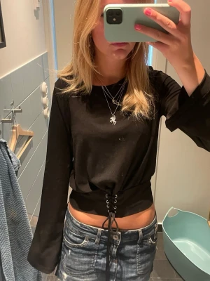 Svart långärmad topp med snörning - Säljer en svart långärmad topp med snörning i midjan. Toppen har en croppad passform och vida ärmar som ger en trendig look. Perfekt att styla med jeans eller kjol för en cool outfit.