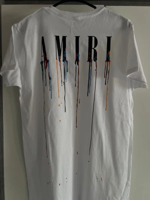 Vit AMIRI t-shirt med färgstänk - Säljer en vit t-shirt från AMIRI med tryck på både fram- och baksidan. Stora bokstäver och färgstänk i blått, rött, orange och svart ger en unik look. Klassisk passform och korta ärmar, perfekt till jeans eller shorts.