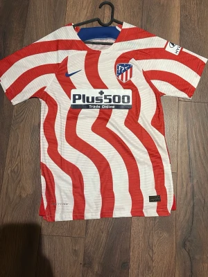 Atletico Madrid spelartröja Nike - Säljer en Atletico Madrid matchtröja från Nike med röda och vita vågiga ränder. Tröjan har klubbmärke på bröstet, blå Nike-logga och sponsortryck på magen. Tillverkad i lätt och ventilerande material, perfekt för fotboll eller träning.