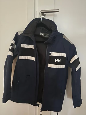 Marinblå Helly Hansen vindjacka - Snygg marinblå vindjacka från Helly Hansen med vita och svarta detaljer. Jackan har hög krage, dragkedja framtill och HH-logga på bröstet, ärmen och nacken. Perfekt för blåsiga dagar och sportiga outfits.