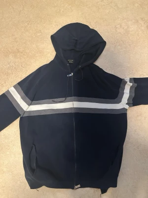 Marinblå hoodie från Mountain Deli - Mörkblå hoodie med dragkedja från Mountain Deli. Tröjan har två breda horisontella ränder i grått och vitt över bröstet och armarna, samt fickor på sidorna och justerbar huva med snören. Materialet är stickat och känns mjukt.