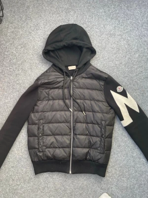 Svart Moncler cardiganjacka med huva - Snygg svart cardiganjacka från Moncler med vadderad framsida, stickade ärmar och stor huva. Ena ärmen har ett vitt zigzag-mönster och Moncler-logga. Jackan har dragkedja framtill och två fickor med dragkedja. Perfekt för dig som vill ha en sportig och trendig look.