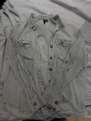 Randig skjorta från Vero Moda - Säljer en snygg vit och grå randig skjorta från Vero Moda. Skjortan har långa ärmar, två bröstfickor med lock och knappar framtill. Perfekt för en avslappnad och stilren look.