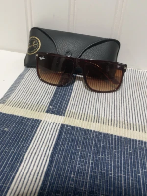 Bruna Ray-Ban solglasögon - Snygga solglasögon från Ray-Ban med rektangulära bågar i mörkbrunt och bruna tonade glas. Klassisk design som passar till alla outfits. Kommer med original Ray-Ban fodral.