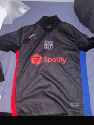 Svart FC Barcelona matchtröja - Säljer en svart FC Barcelona matchtröja med klubbmärke och Spotify-logga på bröstet. Tröjan har röda och blå detaljer på sidorna och en klassisk krage. Perfekt för dig som älskar fotboll och vill visa ditt stöd för Barca.
