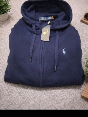 Mörkblå hoodie från Polo Ralph Lauren - Snygg mörkblå hoodie från Polo Ralph Lauren med dragkedja framtill, två fickor och justerbar huva med snören. Klassisk logga broderad på bröstet i ljusblått. Tillverkad i mjukt bomullsmaterial för extra komfort.
