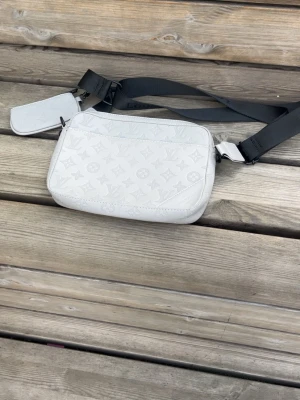Vit axelväska med monogram - Snygg vit axelväska med präglat monogrammönster och bred svart axelrem. Väskan har en rektangulär form och ett extra litet fack på remmen. Perfekt för dig som vill ha en stilren och trendig accessoar till din outfit.