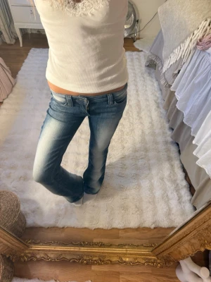 Blå bootcut jeans med slitningar 24/30 - Säljer ett par blå bootcut jeans med snygga slitningar och låg midja. Jeansen har klassiska fem fickor och detaljerade sömmar baktill. Perfekta för en avslappnad och trendig look. Materialet är denim i bomull med lite stretch för extra komfort.