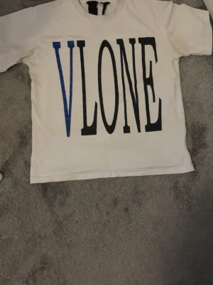 Vit VLONE t-shirt med blått tryck - Säljer en vit t-shirt från VLONE med stort tryck fram i svart och blått. På baksidan finns ett stort blått V med en orm som slingrar sig runt bokstaven. Klassisk passform och tillverkad i mjuk bomull. Perfekt för dig som gillar streetwear och vill sticka ut.