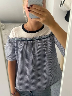 Randig blus med offshoulder-detalj - Säljer en söt blus med tunna blå och vita ränder, offshoulder-detalj och korta ärmar. Överdelen är vit med mörkblå kant vid halsen och resten av toppen är randig. 