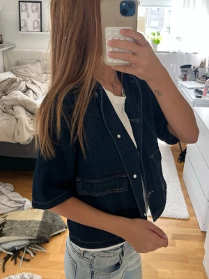 Mörkblå kortärmad jeansjacka - Snygg mörkblå jeansjacka med korta ärmar och vita kontrastsömmar. Jackan har tryckknappar framtill och två stora fickor på sidorna. Perfekt att slänga över en t-shirt för en avslappnad look.
