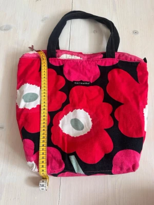 Marimekko Unikko handväska röd - Säljer en ikonisk Marimekko handväska med det klassiska Unikko-mönstret i rött, vitt och svart. Väskan har två svarta handtag och är tillverkad i slitstark bomullscanvas. Perfekt storlek för vardagens essentials och stängs med dragkedja upptill.
