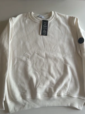 C P tröja  - Klassisk vit sweatshirt med rund hals och ribbade muddar vid ärmslut och nederkant. Tröjan har en diskret patch på ena ärmen och är tillverkad i mjukt material, perfekt för en clean och enkel stil. 100% polyester