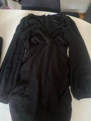 Svart långärmad kortklänning - Snygg svart kortklänning med långa ärmar och v-ringad urringning. Klänningen har en figurnära passform och är tillverkad i ett mjukt, glansigt material som ger en elegant look. Perfekt för dig som vill ha en stilren och trendig outfit.