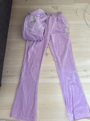 Lila velourset från Juicy Couture - Säljer ett ikoniskt loungewear-set från Juicy Couture i mjuk lila velour. Setet består av en zip-hoodie med huva och stora glittriga Juicy Couture-tryck samt matchande byxor med resår i midjan och logga på benet. Perfekt för chill dagar hemma, de är lite noppriga inifrån men annars helt jätte bra skick.