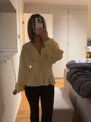 Gul blus med ballongärmar och v-ringning - Trendig gul blus med djup v-ringning och dekorativa knappar framtill. Blusen har långa ballongärmar och markerad midja som ger en snygg siluett. Perfekt att styla med både jeans och kjol för en fräsch look. Storlek 32 men passa mig som bruka ha 34/36. Väldigt stor i storleken. Aldrig använd.