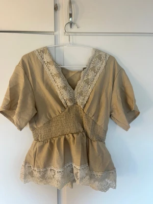 Beige blus med spetsdetaljer - Söt beige blus med korta ärmar och v-ringning dekorerad med vit spets. Blusen har smock i midjan för en feminin siluett och spetskant nertill. Perfekt för dig som gillar romantisk och vintage-inspirerad stil.