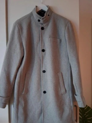 Ljusgrå ullrock med ståkrage. Jack & Jones - Snygg ljusgrå rock i ullblandning med ståkrage och svarta knappar. Rocken har två snedställda fickor fram och en bröstficka. Enkel och stilren design som passar perfekt till höst och vinter.