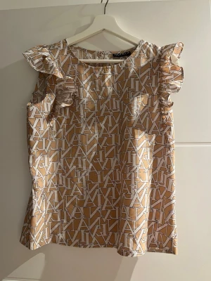Beige blus med volangärmar och mönster - Trendig beige blus med vitt grafiskt mönster och volangdetaljer vid axlarna. Blusen är ärmlös och har en rund halsringning. Materialet ser ut att vara lätt och glansigt, perfekt för en snygg och modern look.