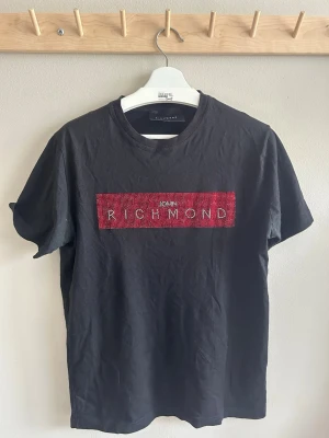 Svart Richmond t-shirt med röd detalj - Säljer en svart t-shirt från Richmond med en stor röd detalj framtill där märket står i vita bokstäver. T-shirten har korta ärmar och är gjord i 100% bomull. Perfekt för dig som gillar en enkel men ändå statement look. Köpte den på Zgander i Helsingborg. Inget kvitto kvar. 