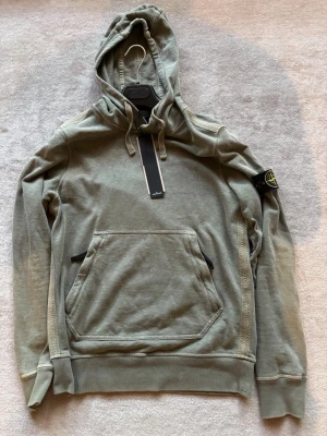 Grön hoodie från Stone Island - Säljer en grön hoodie från Stone Island med klassisk patch på vänster ärm. Hoodien har stor känguruficka framtill, dragsko i huvan och ribbade muddar. Materialet är mjukt och bekvämt, perfekt för en avslappnad streetwear-look.