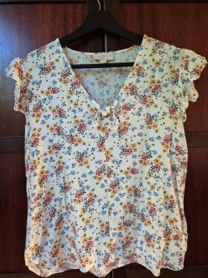 Blommig blus - Söt blommig blus i vitt med små blommor i rosa, blått och gult från Loft. Blusen har v-ringning med knyt framtill och korta volangärmar. Perfekt för vår och sommar, till jeans eller kjol.