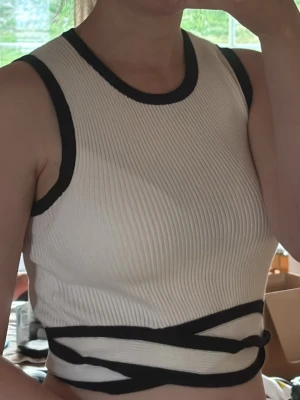 Vit ribbad croppad linnetopp - Säljer en vit ribbad croppad linnetopp med svarta kontrastkanter och snygga banddetaljer i midjan. Toppen är ärmlös och har rund halsringning. Perfekt för dig som vill ha en stilren men ändå unik topp till sommaren. Toppen är oanvänd men prislappen har åkt av!