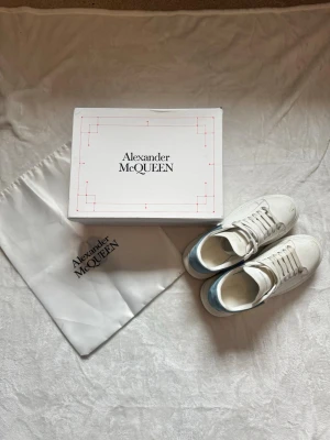 Vita Alexander McQueen sneakers - Säljer ett par klassiska vita sneakers från Alexander McQueen med blå detalj på hälen. Skorna har tjock sula, rund tå och breda vita skosnören. Tillverkade i läder och kommer med originalkartong och dustbag.